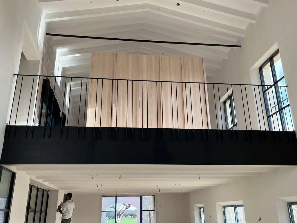 Interior de una casa moderna en construcción con doble altura, ventanas grandes, estructura de techos expuestos y un mezzanine con barandal de metal.