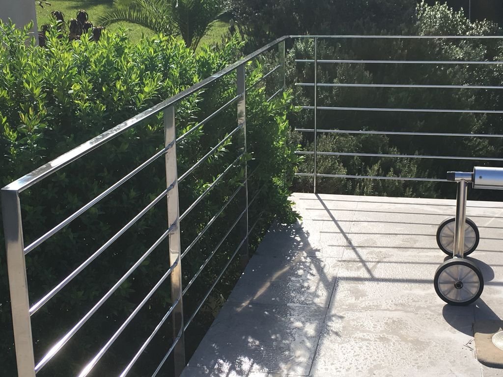 Barandal de acero inoxidable en un balcón o terraza con sombra de plantas y árboles cercanos.