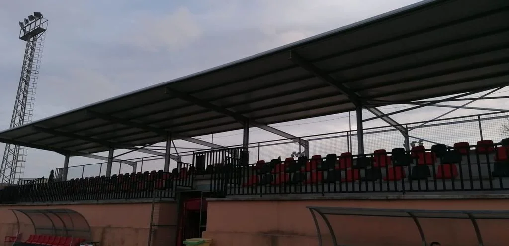 Gradas de un estadio cubiertas con asientos rojos y negros, con una valla y estructura metálica, bajo un cielo nublado.