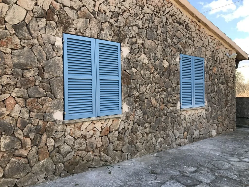 Fachada de una casa de piedra con dos ventanas de persianas azules y un suelo de piedra.