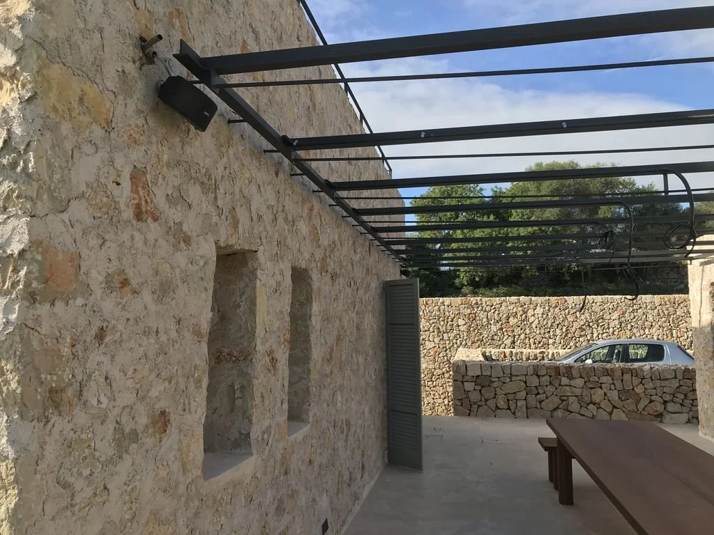 Pared de piedra con ventanas, estructura de metal y coche estacionado en el fondo.