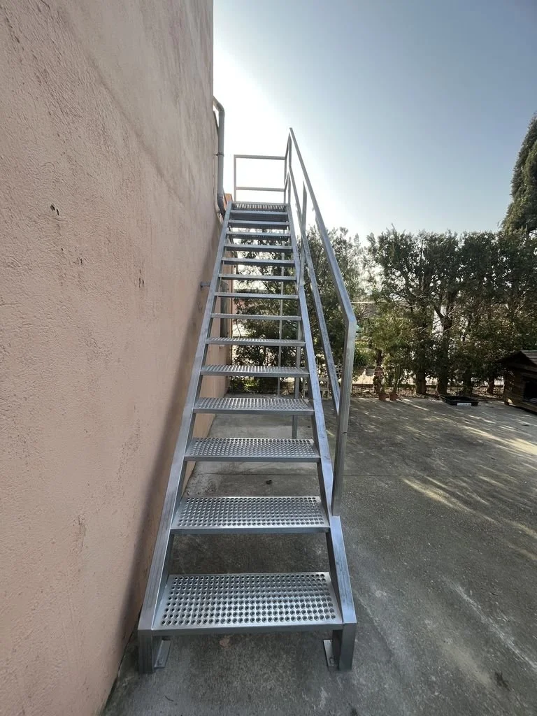 Escalera metálica exterior junto a una pared de color beige, con barandillas a ambos lados, en un espacio al aire libre con árboles y suelo de concreto.