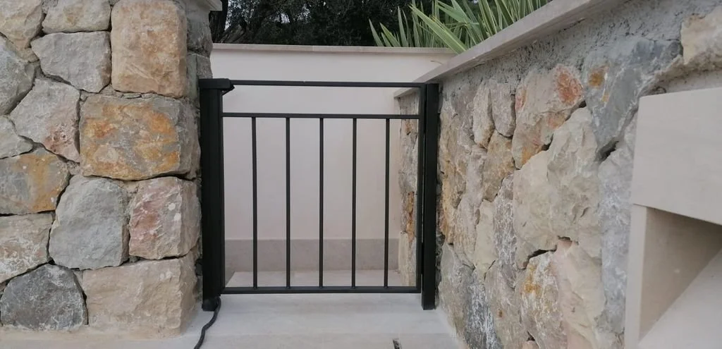 Puerta pequeña de reja negra entre dos muros de piedra en un espacio exterior.