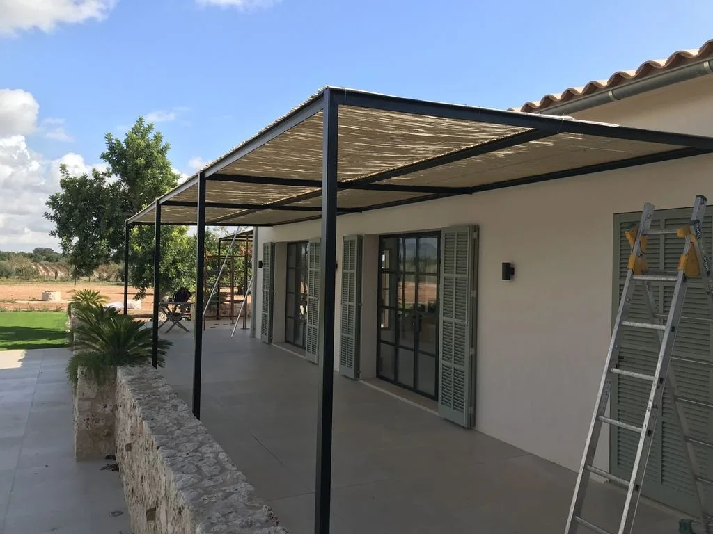 Terraza con estructura metálica y toldo de láminas, puerta de cristal con persianas, escaleras, plantas y paisaje exterior con árboles y campo.