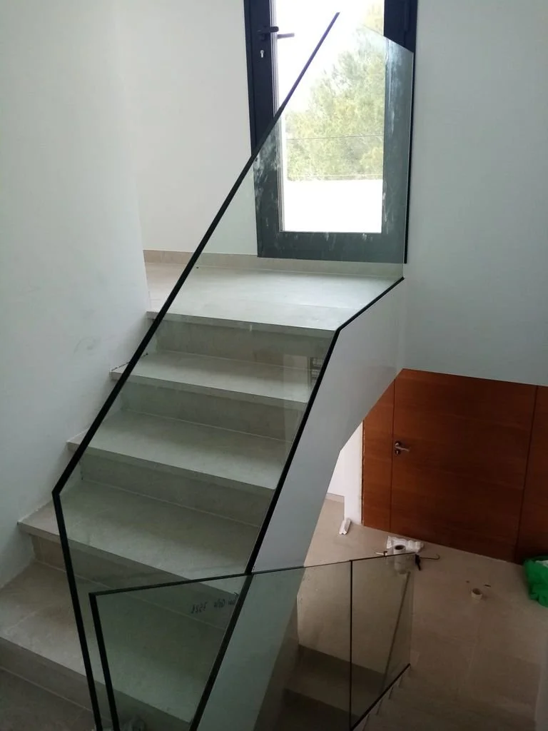 Escalera de cristal con barandilla negra, piso de mármol, ventanas y paredes blancas en un espacio interior.