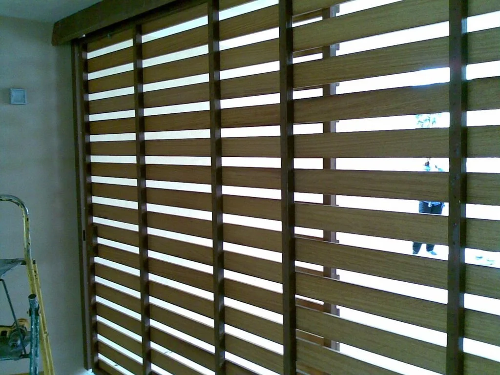 Puerta o pared de madera con ranuras horizontales, parcialmente abierta, en un interior con una escalera y una pared a la izquierda. Se puede ver a personas y un árbol en el exterior.
