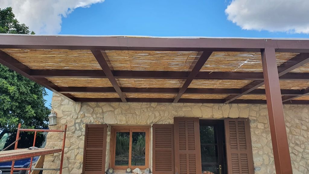 Vista de una casa de piedra con ventanas de madera y persianas, con un porche de madera con estructura de acero y techo de cañas, en un día soleado con cielo azul y algunas nubes.