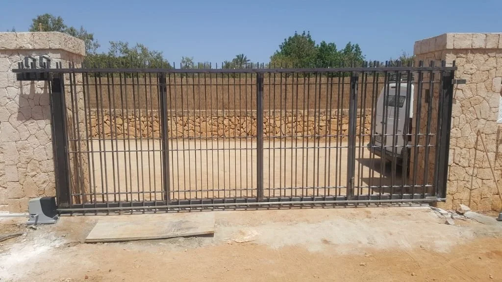 Puerta de reja metálica en la entrada de un muro de piedra, con una camioneta estacionada detrás.