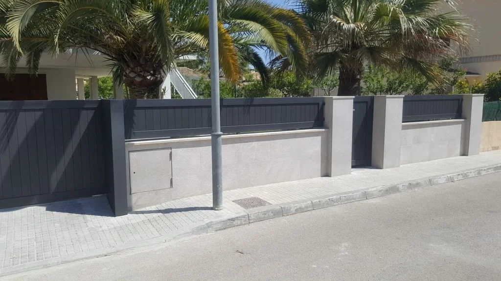 Valla de concreto oscuro con paneles de madera en la parte superior y una puerta, junto a una farola y palmeras en el fondo.