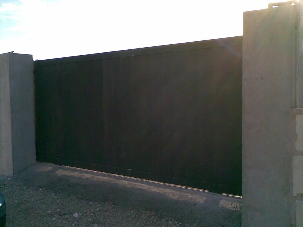 Puerta de metal oscura en un muro de concreto, con sol brillante en el cielo.