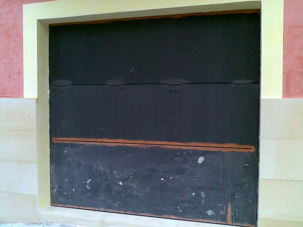 Puerta de garaje de color negro en un muro con paredes de diferentes colores, rosa y beige, en un edificio.
