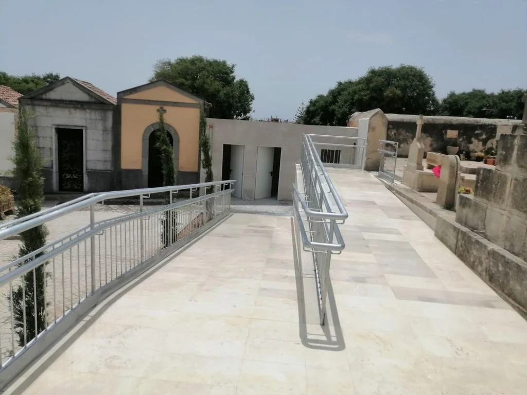 Pasillo de acceso con rampa de metal en un cementerio, con mausoleos y árboles al fondo.