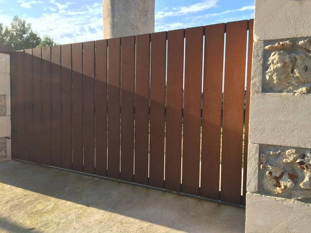 Puerta de madera de listones en color marrón, con muro de piedra a los lados, en exterior con cielo despejado y árboles al fondo.