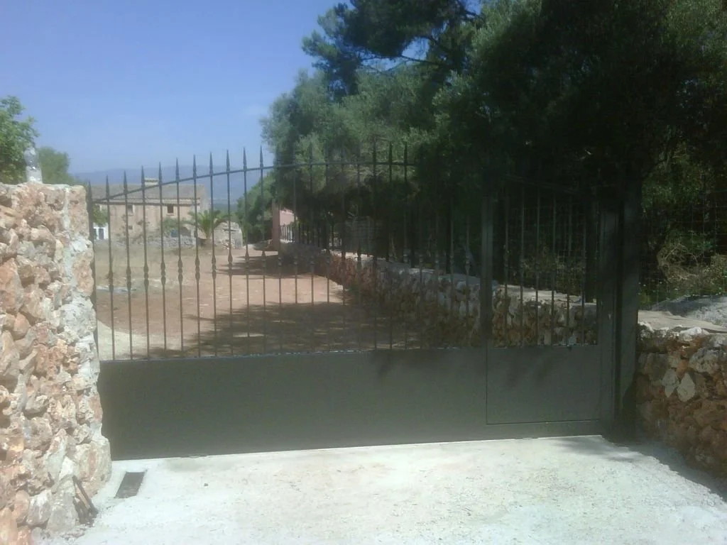Puerta negra de reja con muro de piedra a los lados, en un entorno natural con árboles y un camino de tierra.