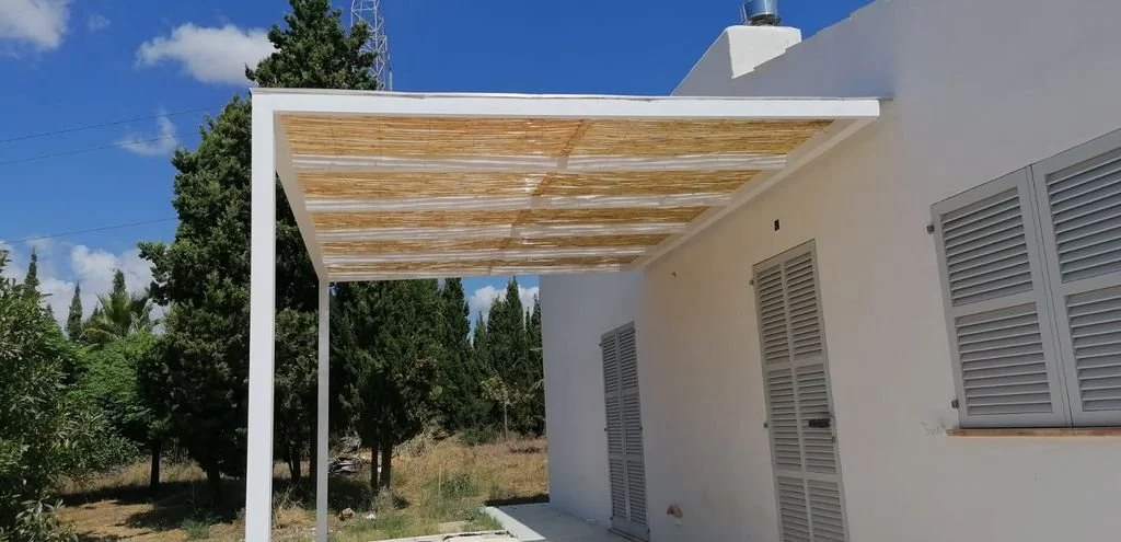 Porche de una casa blanca con techo de madera, en un entorno rodeado de árboles y cielo azul con nubes.