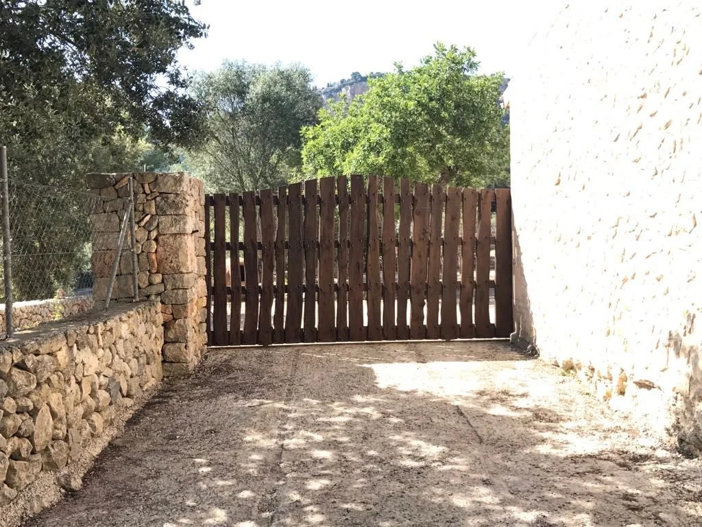 Puerta de madera en un camino de tierra, con muro de piedra a la izquierda y muro de piedra clara a la derecha, rodeada de árboles verdes y cielo despejado.