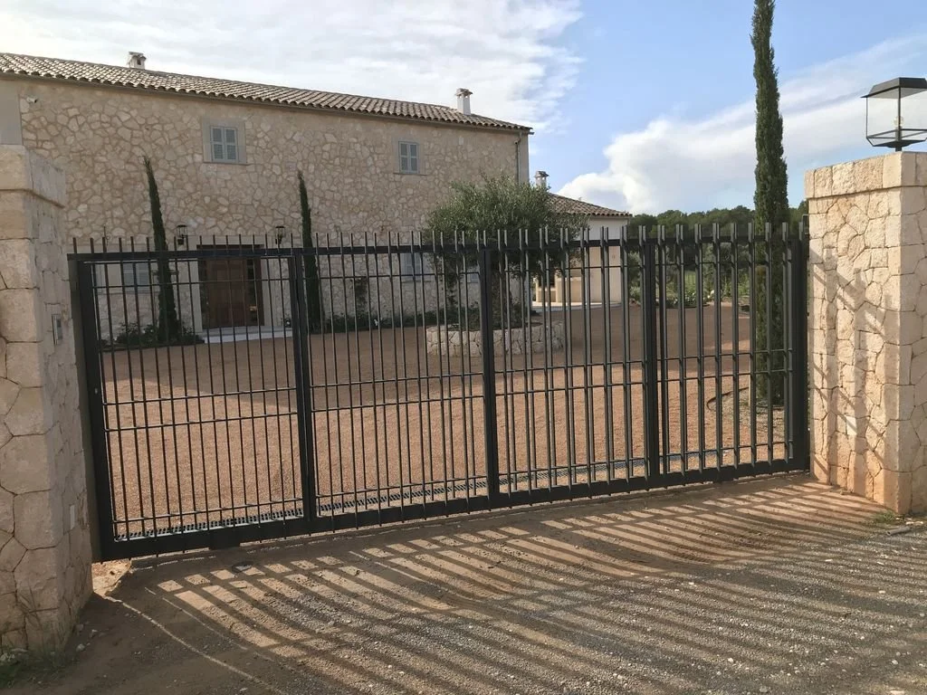 Vista de una verja de barrotes negros que cierra una entrada a una propiedad con una casa de piedra y árboles detrás.