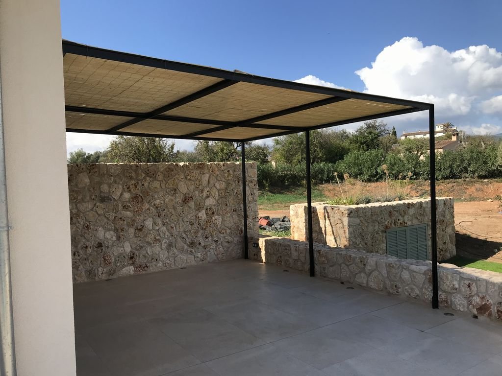 Espacio exterior con toldo de sombra, paredes de piedra y paisaje de naturaleza con árboles y casas al fondo.