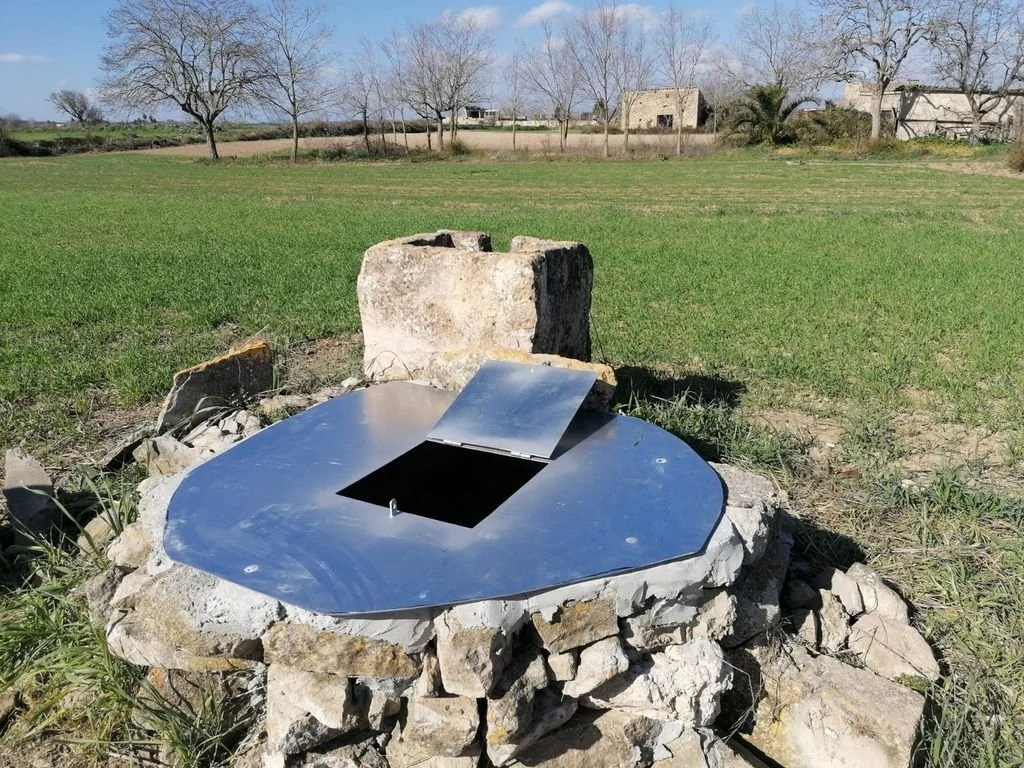 Pozo con tapa metálica en un campo con árboles y edificios en el fondo.