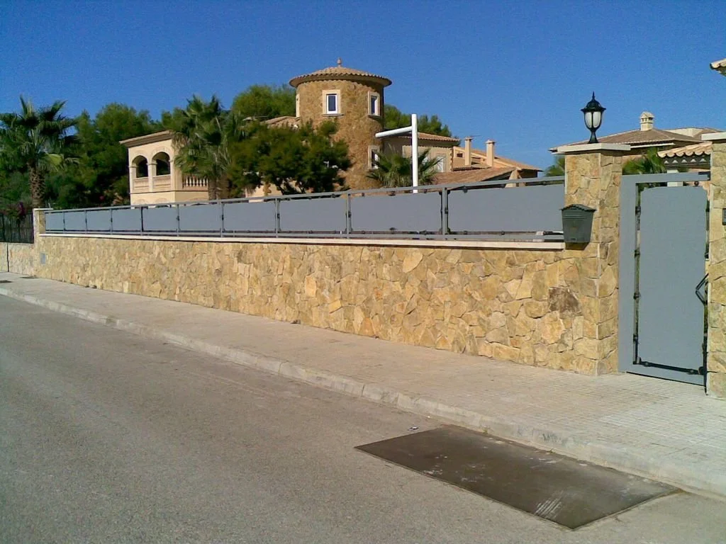 Valla de piedra con cerca metálica y puerta de hierro, en un vecindario con casa grande y palmeras, en un día soleado.