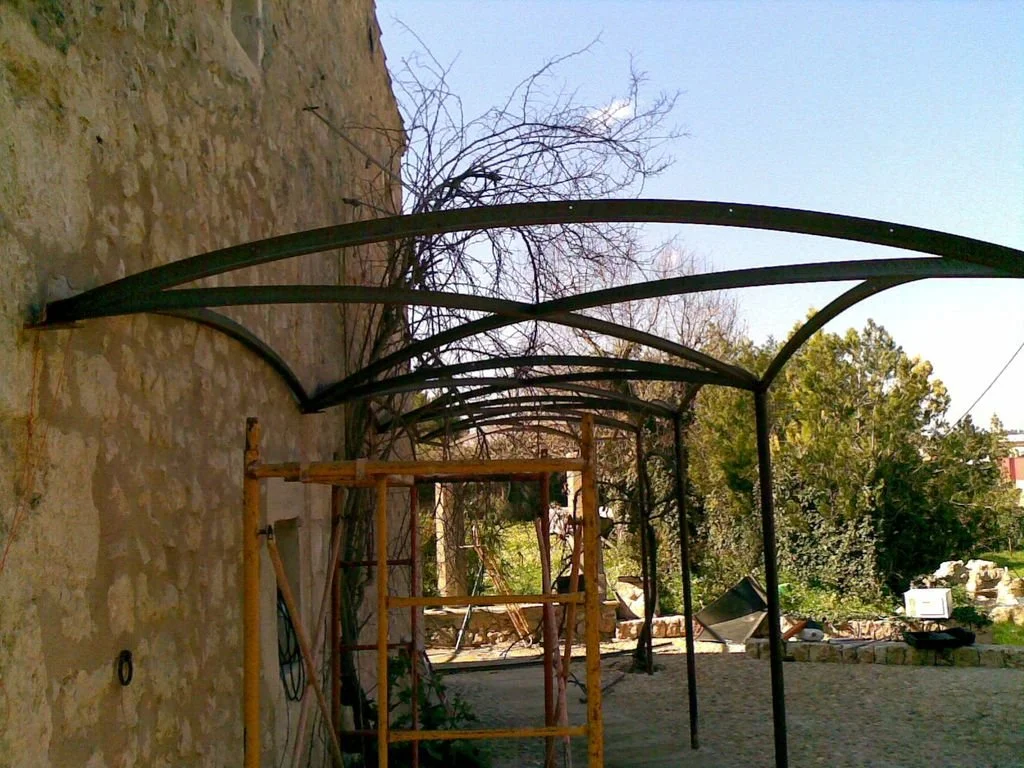 Estructura metálica en construcción junto a un muro de piedra, con una escalera de madera y un fondo con árboles y cielo despejado.