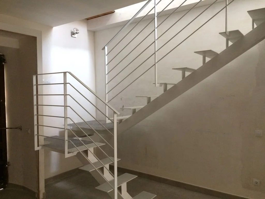 Escalera interior de metal pintada de blanco en un espacio moderno con paredes blancas y iluminación natural.
