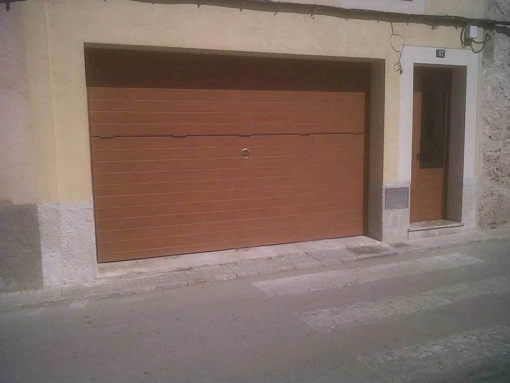 Vista de un garaje con puerta de madera color marrón y acceso peatonal a un lado, en una calle de una zona residencial.