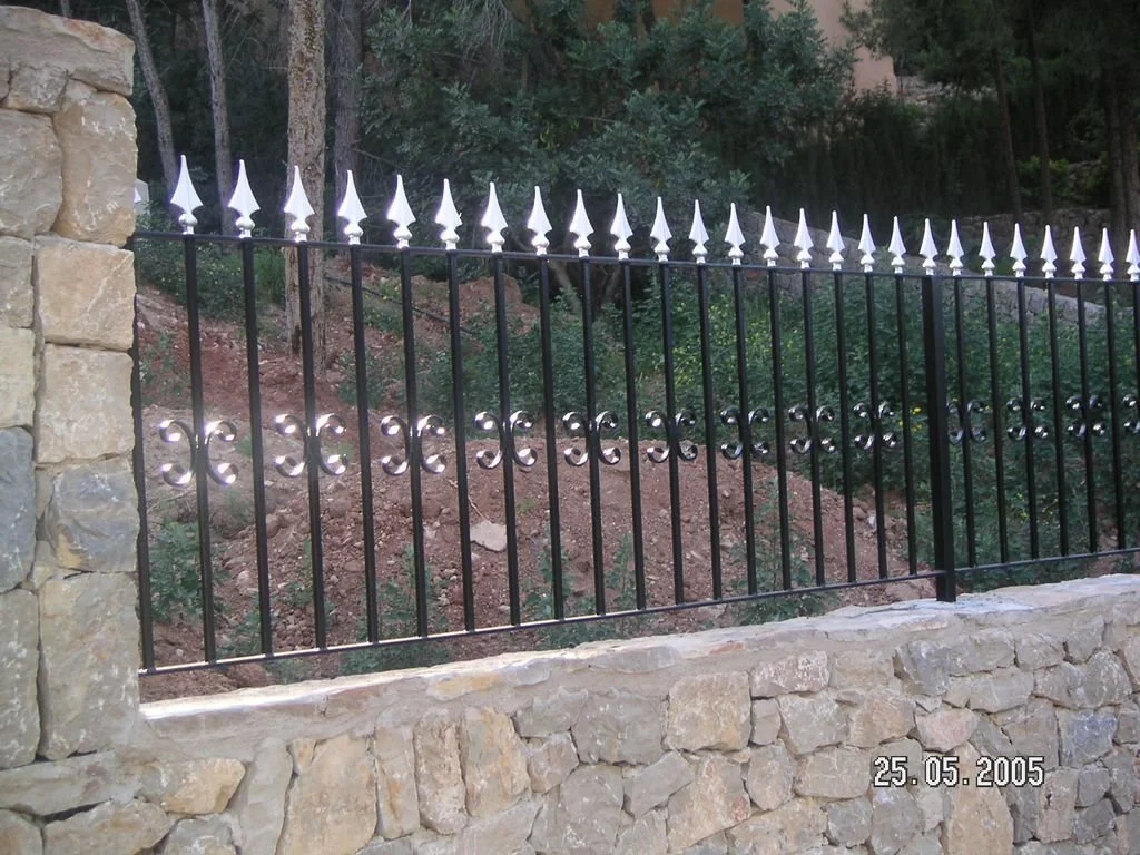 Valla de hierro negro con puntas decorativas blancas en un muro de piedra, en un entorno natural con árboles y vegetación.