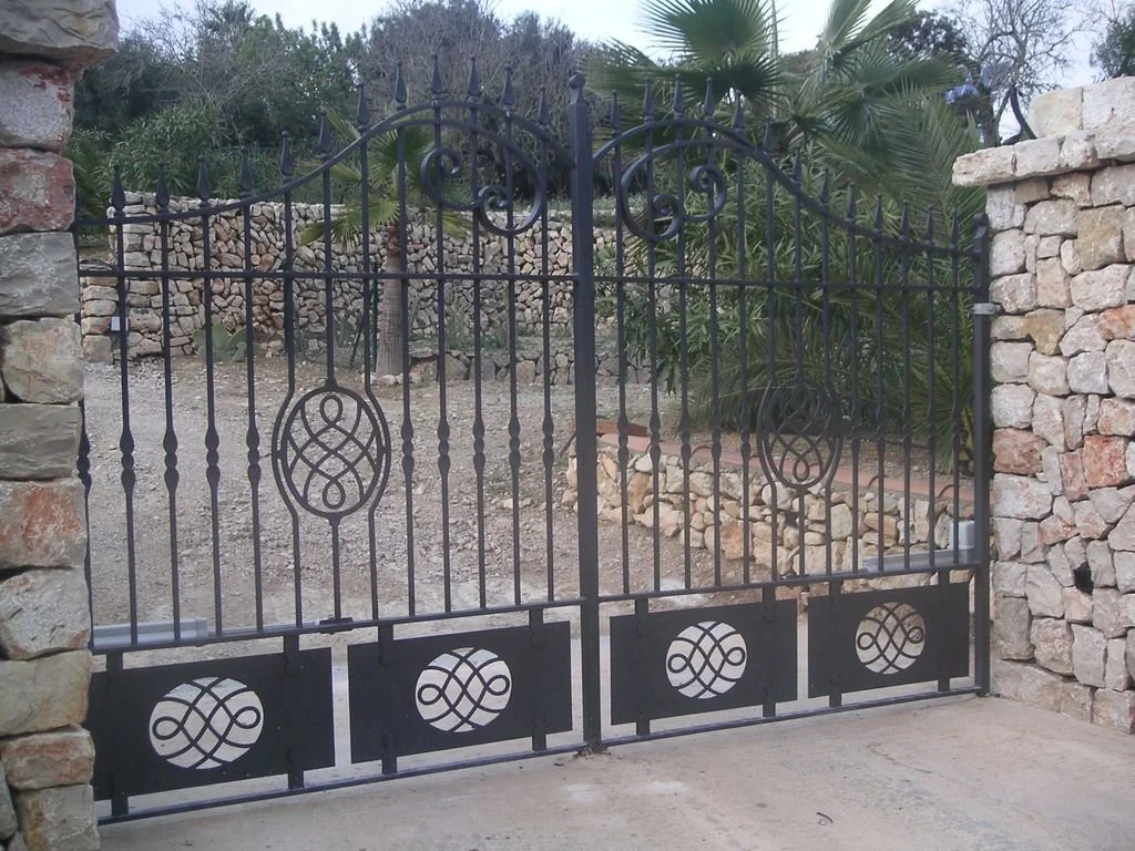 Puerta de reja negra decorativa en un muro de piedras, con vegetación de arbustos y palmeras en el fondo.