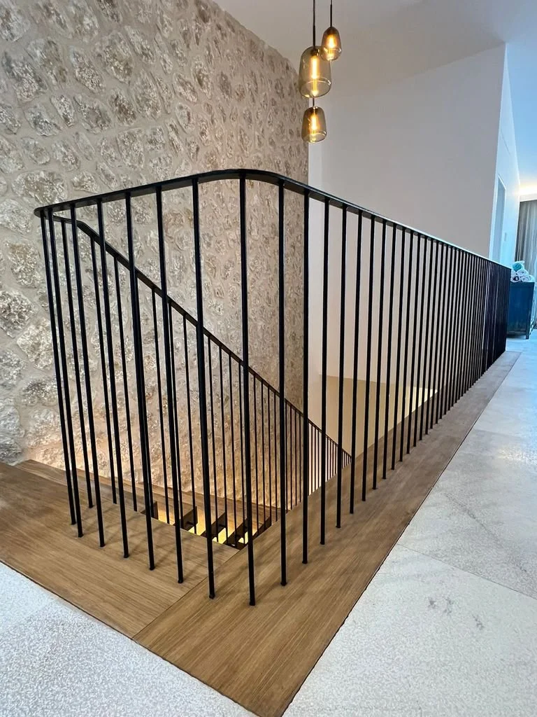 Barandal negro de hierro que rodea una escalera en un espacio interior con pared de piedra y lámparas colgantes doradas.