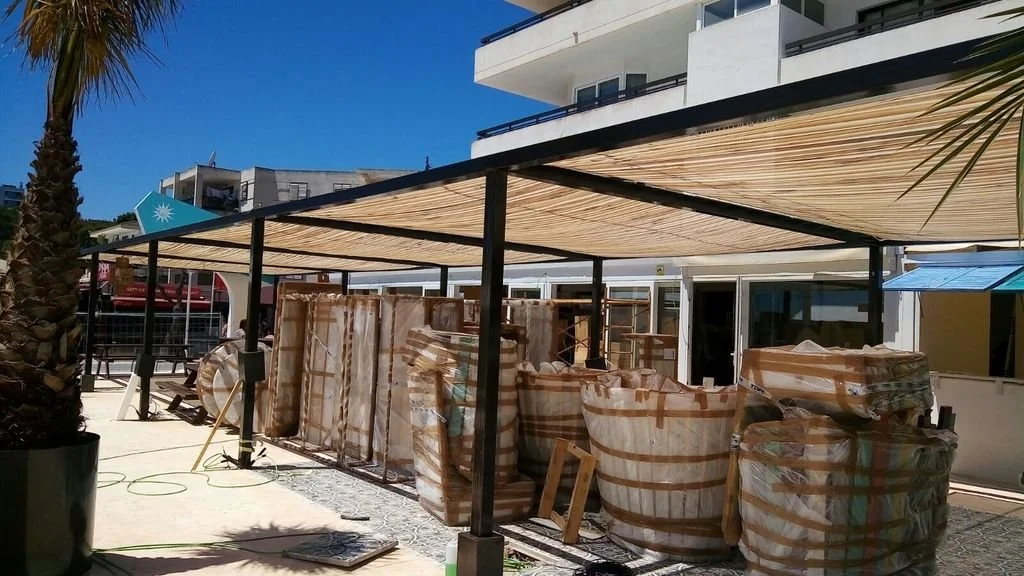 Montón de cestas y tarros grandes apilados en un área exterior con toldo y edificios modernos al fondo.