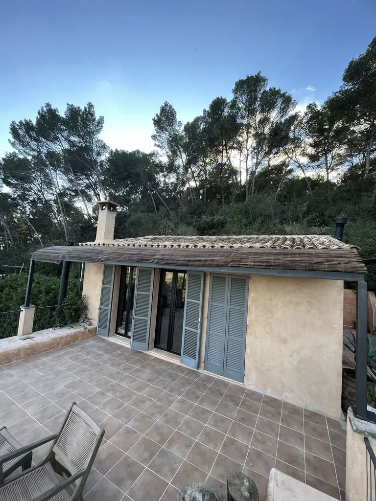 Casa de estilo rústico con ventanas de persianas de madera en un área de terraza con sillas de mimbre, rodeada de árboles y un cielo despejado.