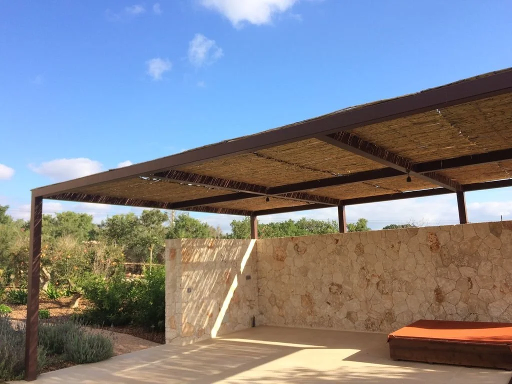 Área de descanso con techo de material aislante, pared de piedra y un sillón en un lugar al aire libre con vegetación y cielo azul.