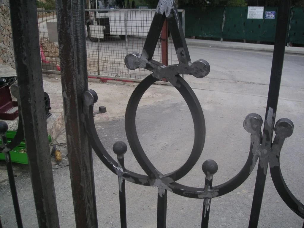 Detalle de reja de metal adornada con elementos circulares y cruz, en un entorno al aire libre con una carretera y vehículos de fondo.