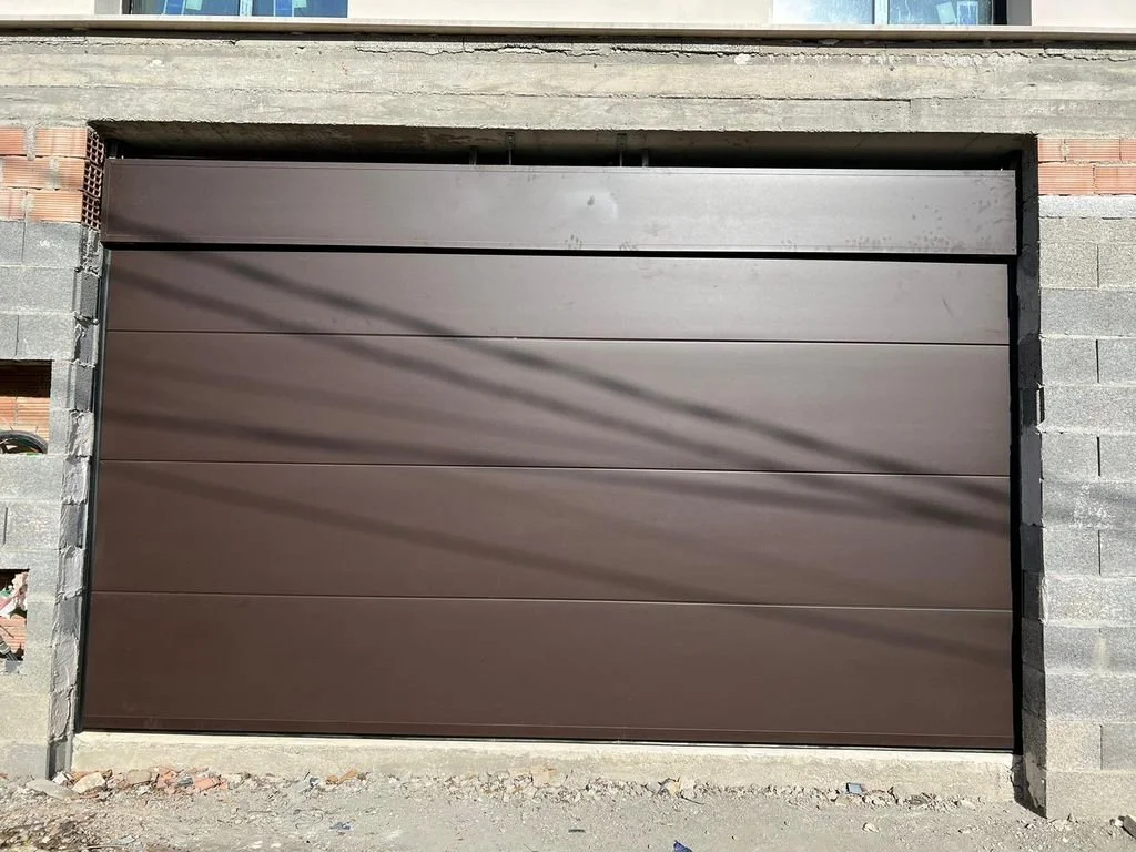 Puerta de garaje metálica de color marrón en construcción, paredes de bloques de cemento y ladrillos visibles alrededor.