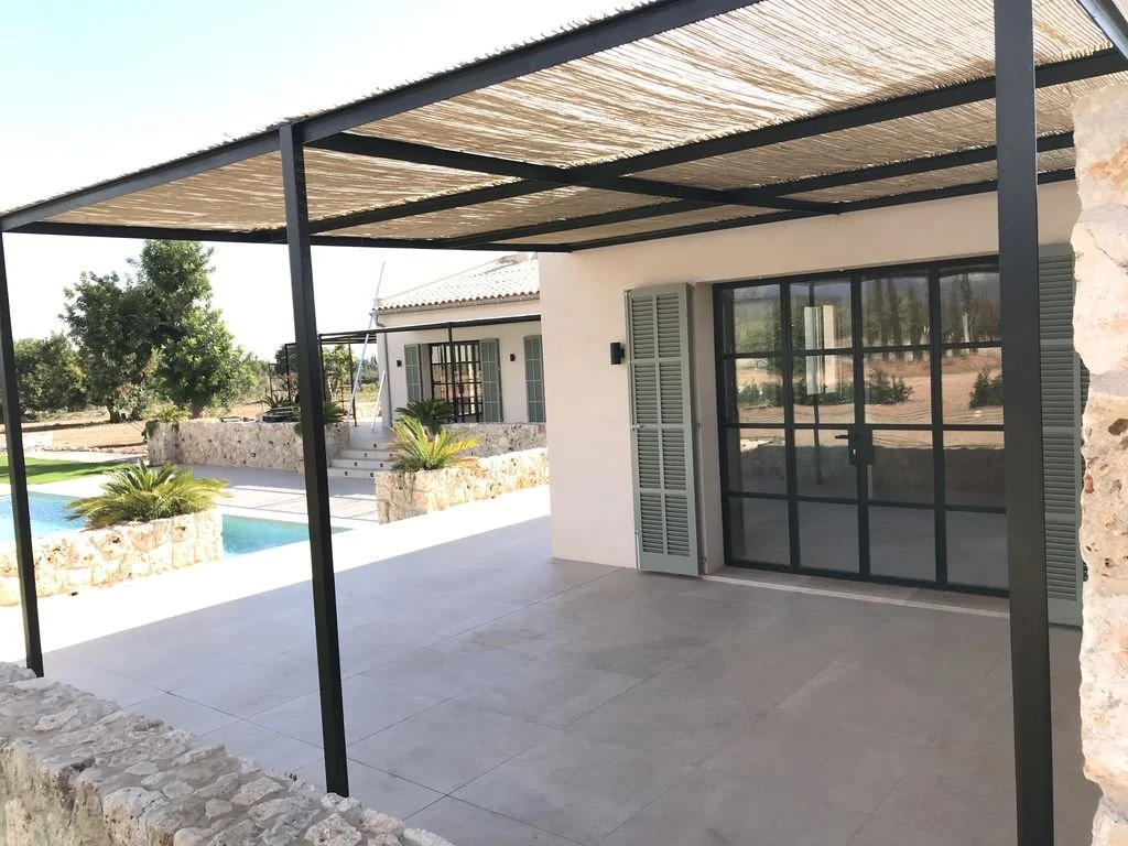 Patio con toldo de caña, piso de baldosas, ventana de cristal con marco negro y cierra de madera gris, zona de jardín con piscina y plantas en macetas de piedra, en un entorno soleado y árido.