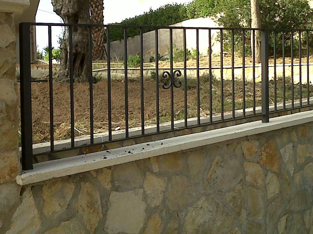 Barandal de metal negro instalado en una ventana, con una pared de piedra en la parte inferior y un entorno al aire libre con árboles y tierra en el fondo.