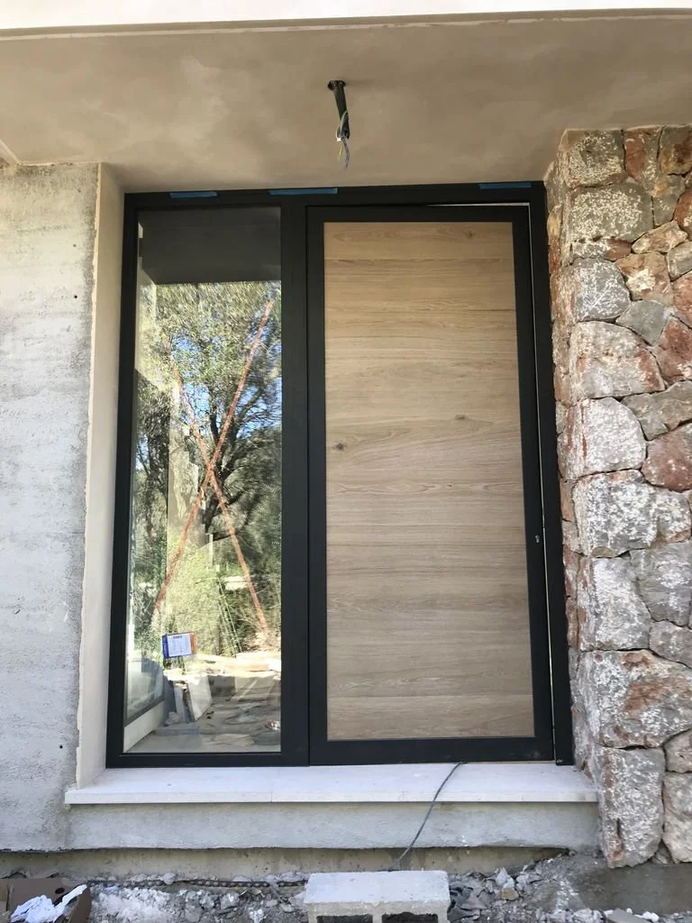 Ventana grande de aluminio negro con una parte de cristal y otra de madera en un muro en construcción.