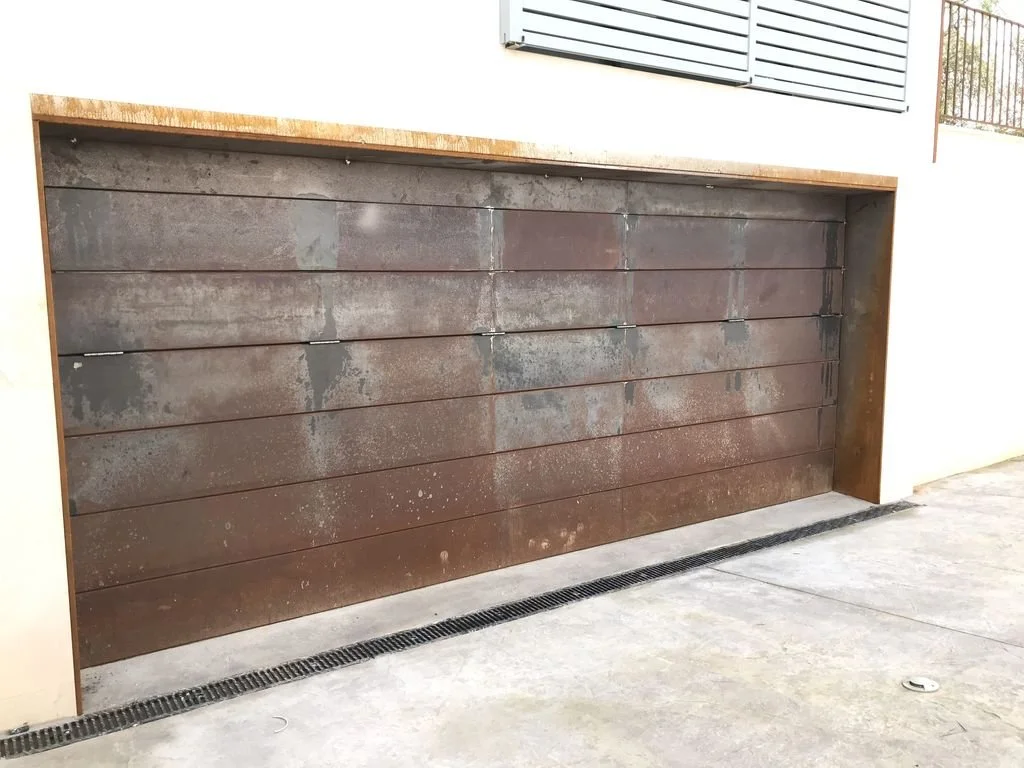Puerta de garaje de madera envejecida con manchas de óxido y pintada de color marrón, instalada en una estructura de color beige.