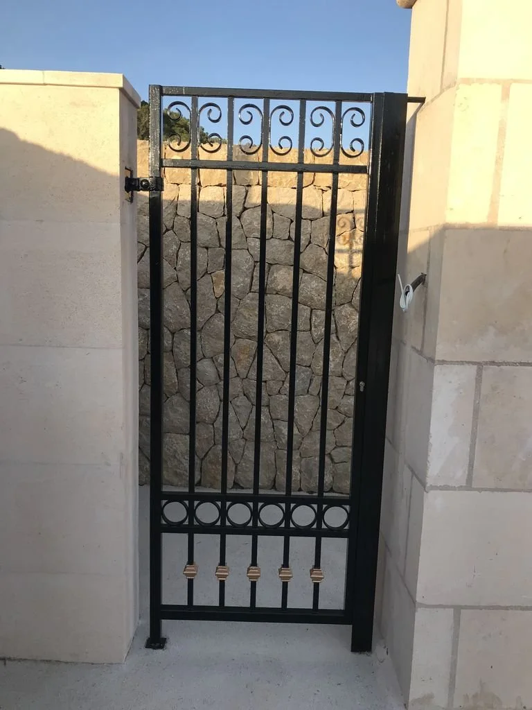 Puerta negra de metal con diseño decorativo en la parte superior, instalada entre muros de piedra y bloques claros, con vista a un muro de piedra en el fondo.