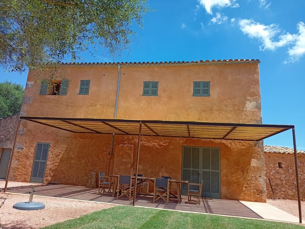 Casa de dos pisos con paredes de ladrillo tostado, ventanas con contraventanas grises, terraza con toldo y muebles de madera, en un día soleado con cielo azul y algunas nubes.