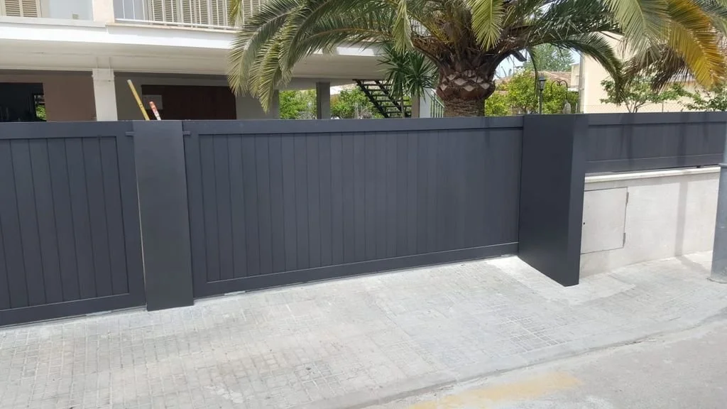 Una cerca de madera gris junto a una acera en una zona urbana con árboles y un edificio de fondo.
