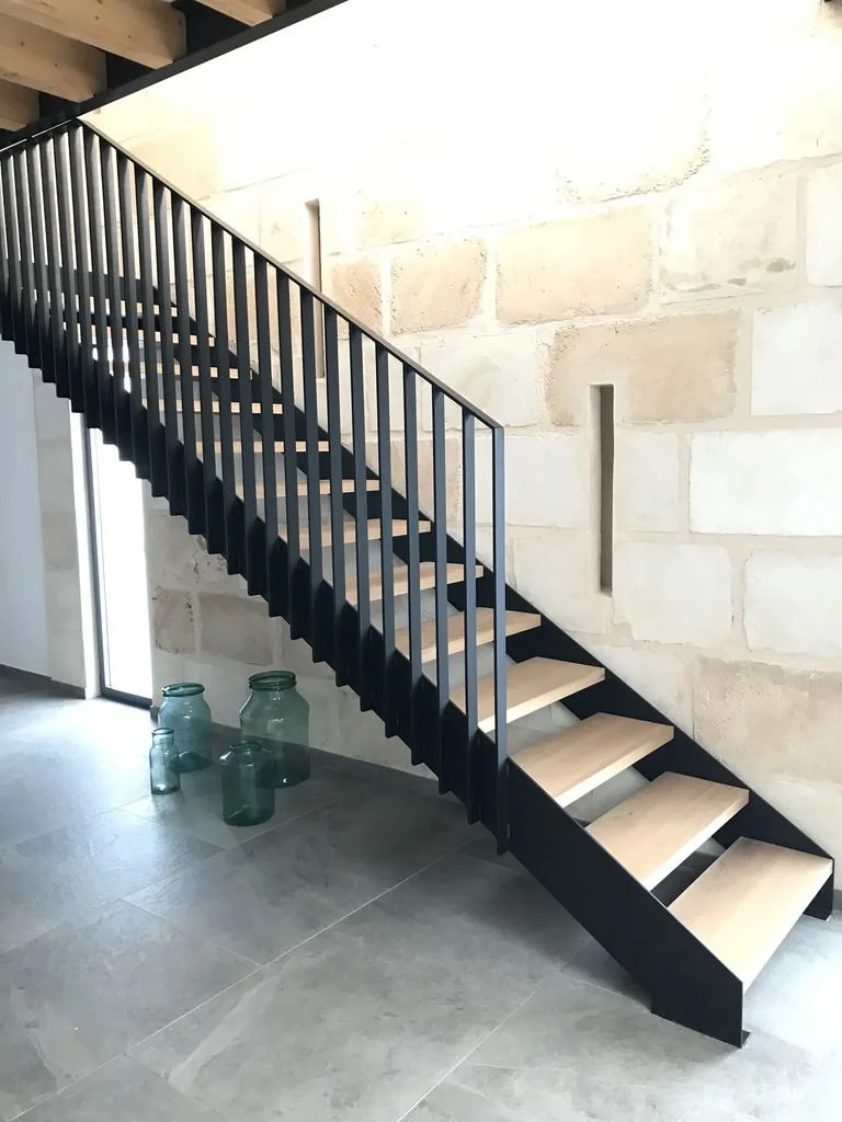 Escalera moderna de madera y metal en interior con paredes de piedra y jarras de vidrio en el suelo.