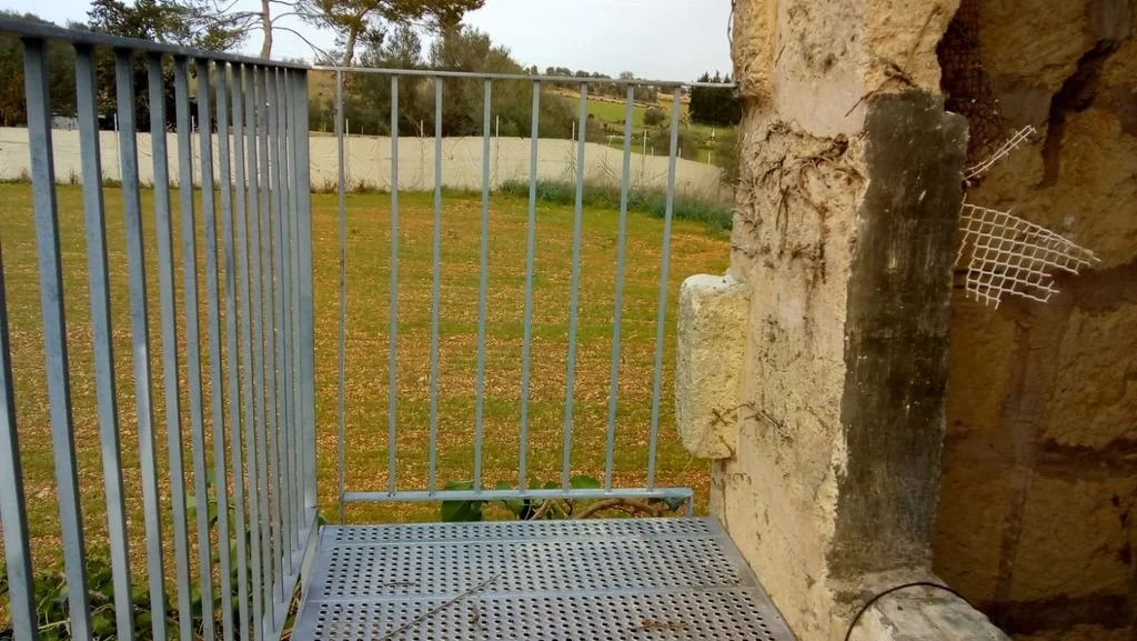 Vista desde un balcón pequeño con barandilla de metal, bordea un muro de piedra con grietas y restos de malla metálica. En el fondo se observa un área de césped y árboles a lo lejos.