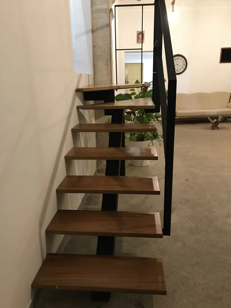 Escalera moderna de madera con barandal negro en un ambiente de sala con sillón blanco y plantas