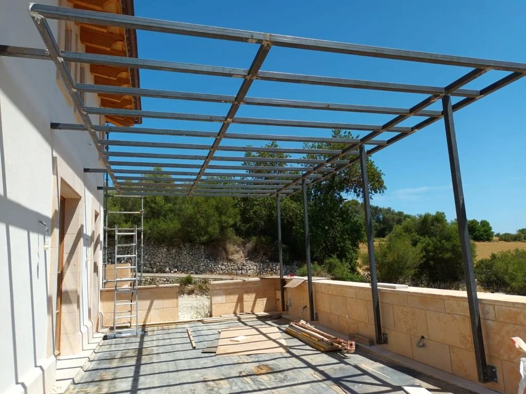 Estructura metálica en construcción en una terraza de una casa, con un cielo despejado y árboles verdes en el fondo.