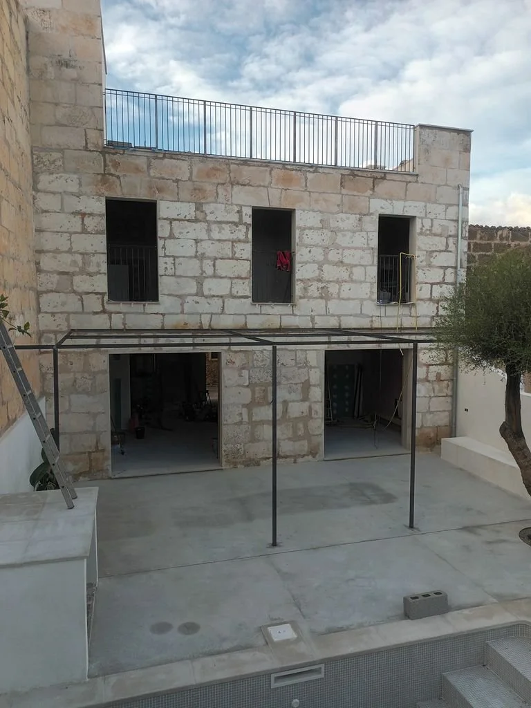 Construcción de una casa con paredes de piedra, ventanas sin marcos y un marco metálico en el patio, con una piscina y un árbol a la derecha.