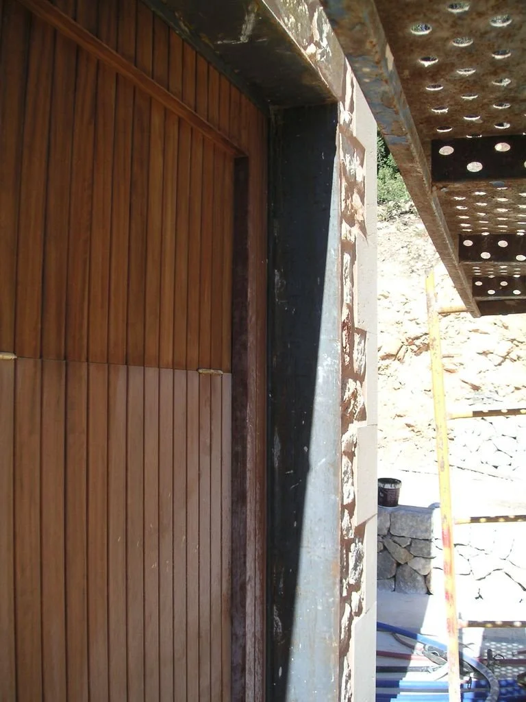 Puerta de madera enmarcada por estructura de concreto y acero, en construcción, con escaleras de andamio al fondo y rocas en un ambiente exterior.