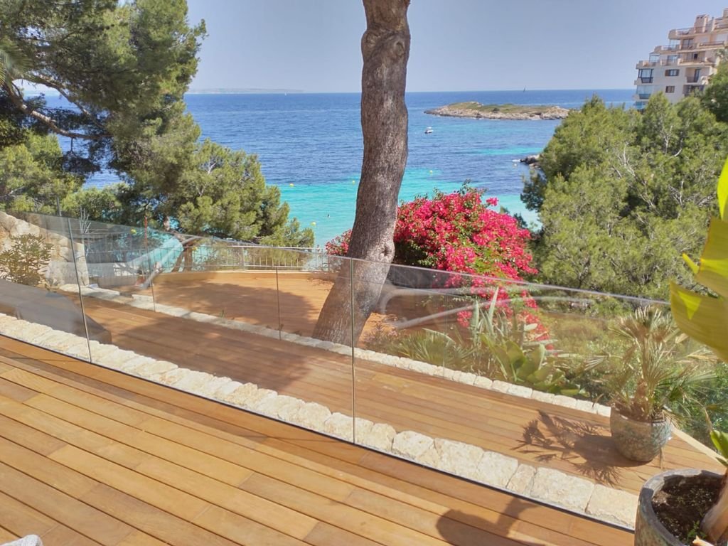 Vista desde una terraza de madera con barandal de vidrio, árboles y plantas tropicales, incluyendo buganvilia rosa, con vista al mar, rocas y un pequeño islote en la distancia, en un día soleado.