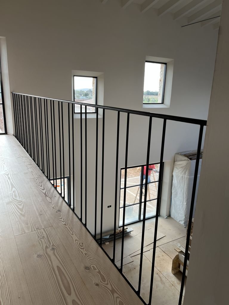 Interior de una casa en construcción con una barandilla de metal negro en un segundo piso, paredes blancas y ventanas que dejan ver el exterior.